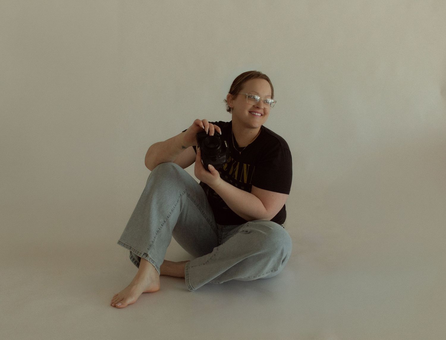 Person sitting cross-legged on floor in black tee and gray jeans, holding a camera, smiling, wearing glasses.