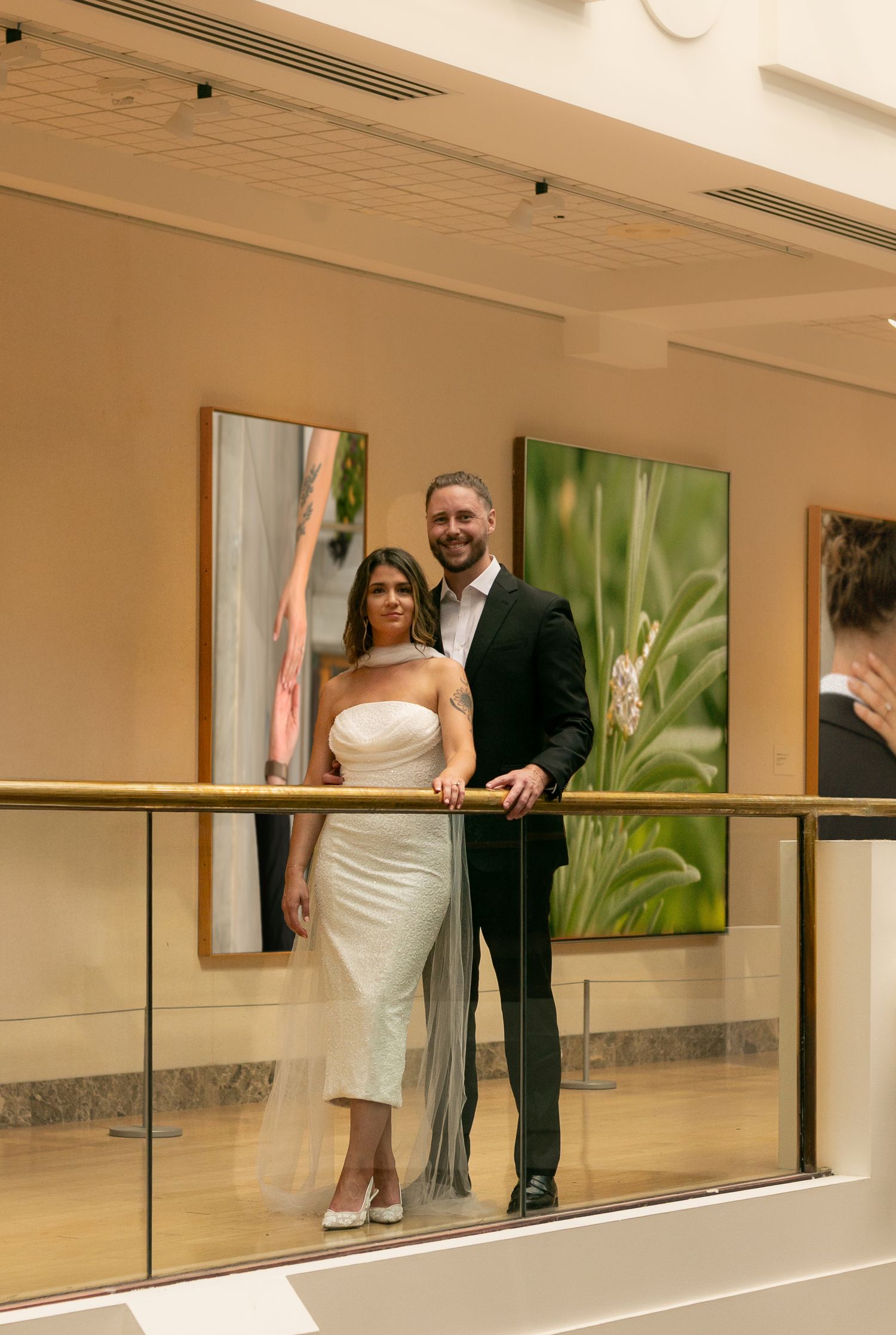 Elegant couple in wedding attire posing on a balcony with gold railing and green botanical artwork behind them.