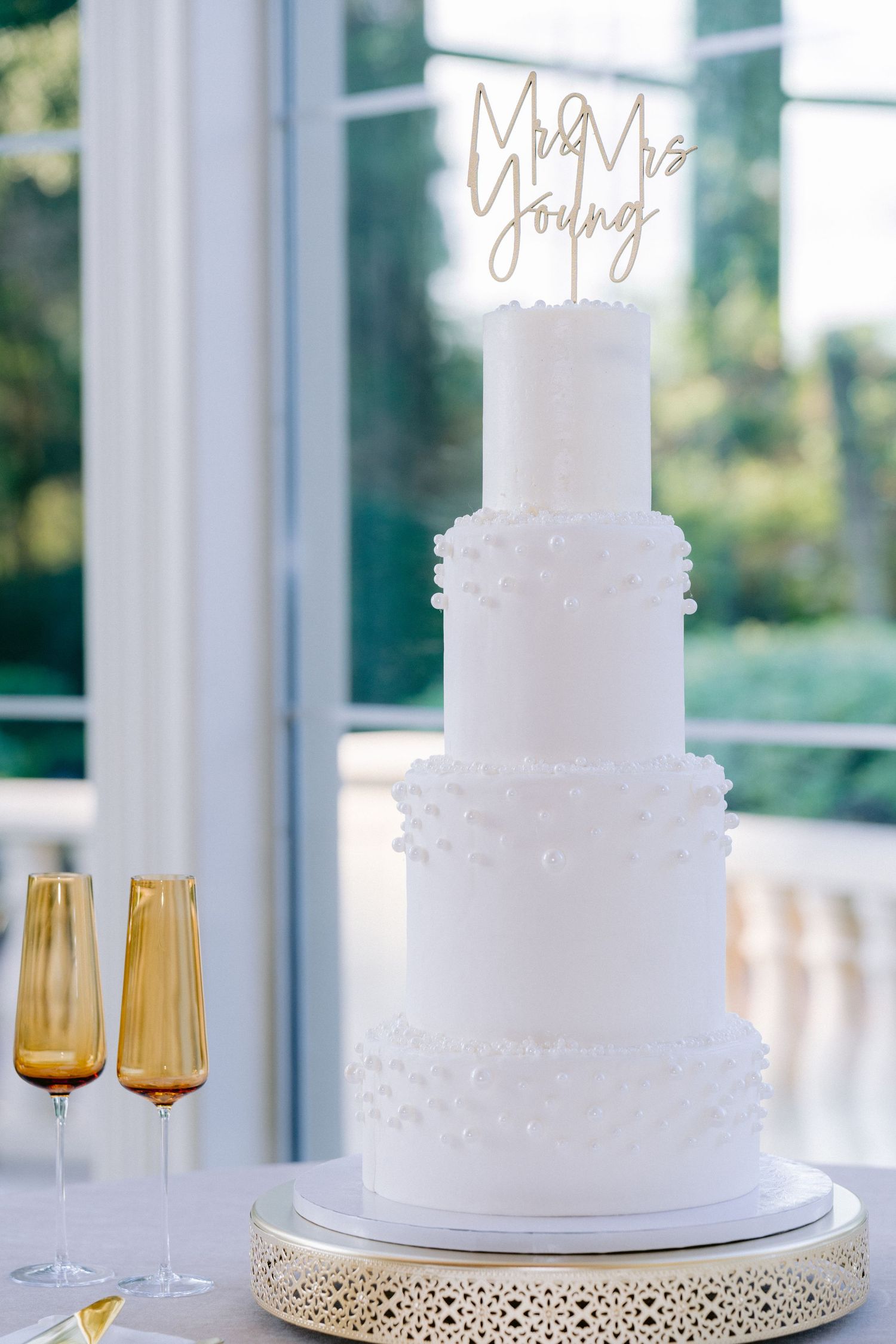 Three white wedding cakes on gold stands with amber glassware displayed in front of bright windows. Tampa Bay Wedding Photographer