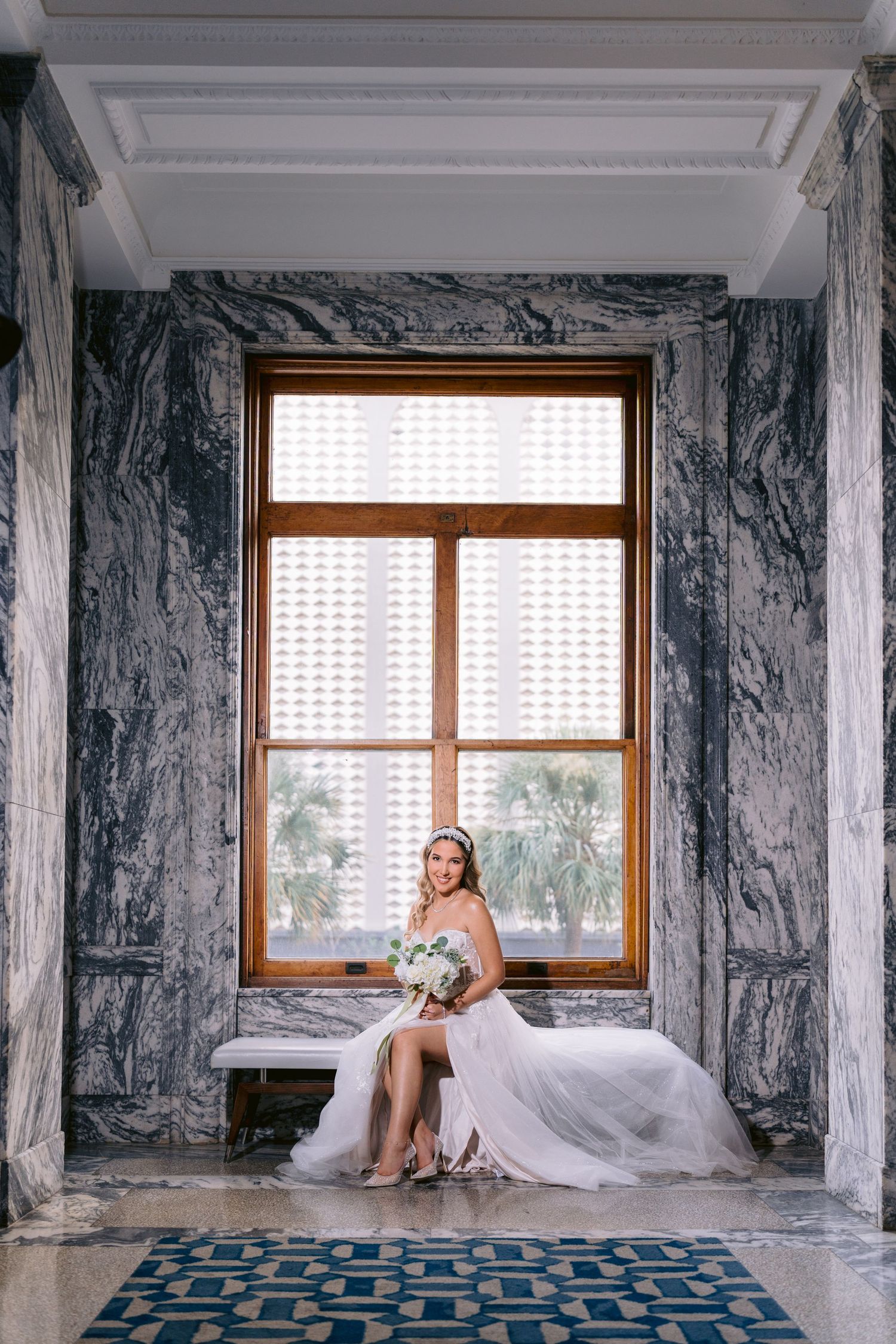 A bride in a white gown rests elegantly against marble walls next to large windows in an opulent hotel setting. Tampa Wedding Photographer