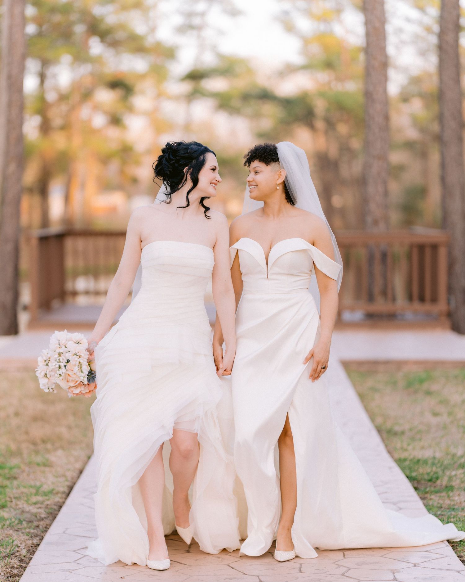 Two brides in elegant white wedding gowns hold hands while walking on a wooden deck surrounded by autumn trees. Tampa Wedding Photographer