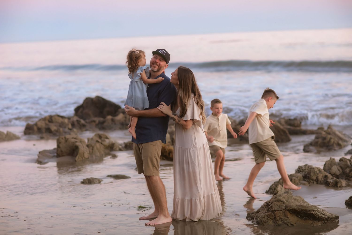 A family enjoys quality time together on a rocky beach at sunset with ocean waves in the background.