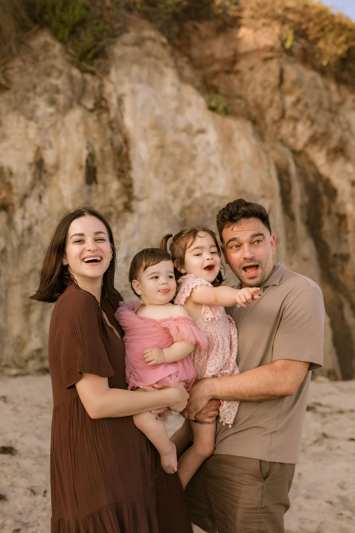 A family shares joyful moments together on a beach with rocky cliffs in the background during a photo session.