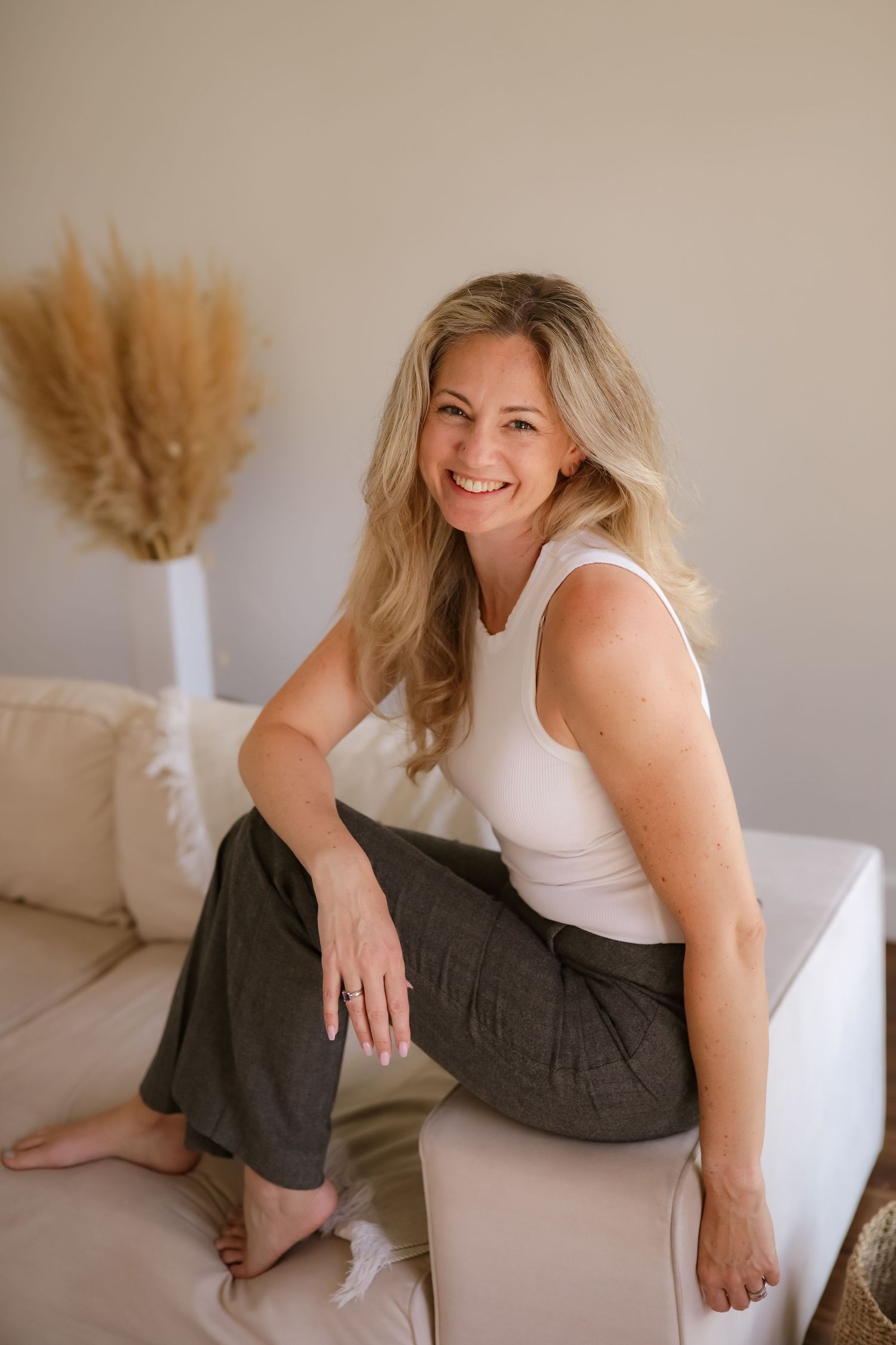 A woman in a white tank top and dark pants sits casually on a beige couch with pampas grass decor in the background.