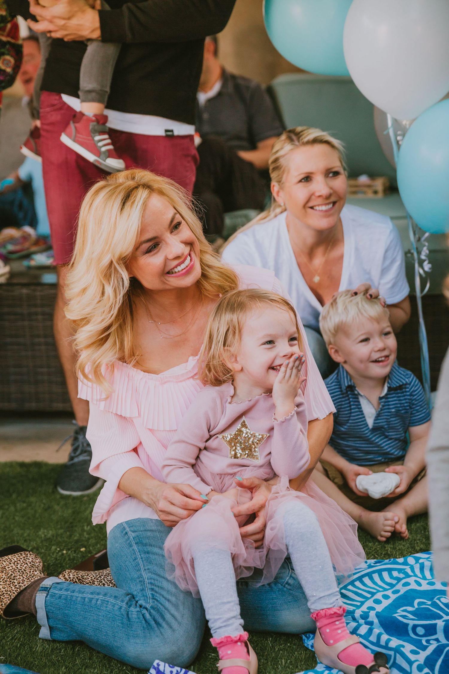 Family enjoying outdoor celebration with blue balloons and blanket while sitting on grass and laughing together.