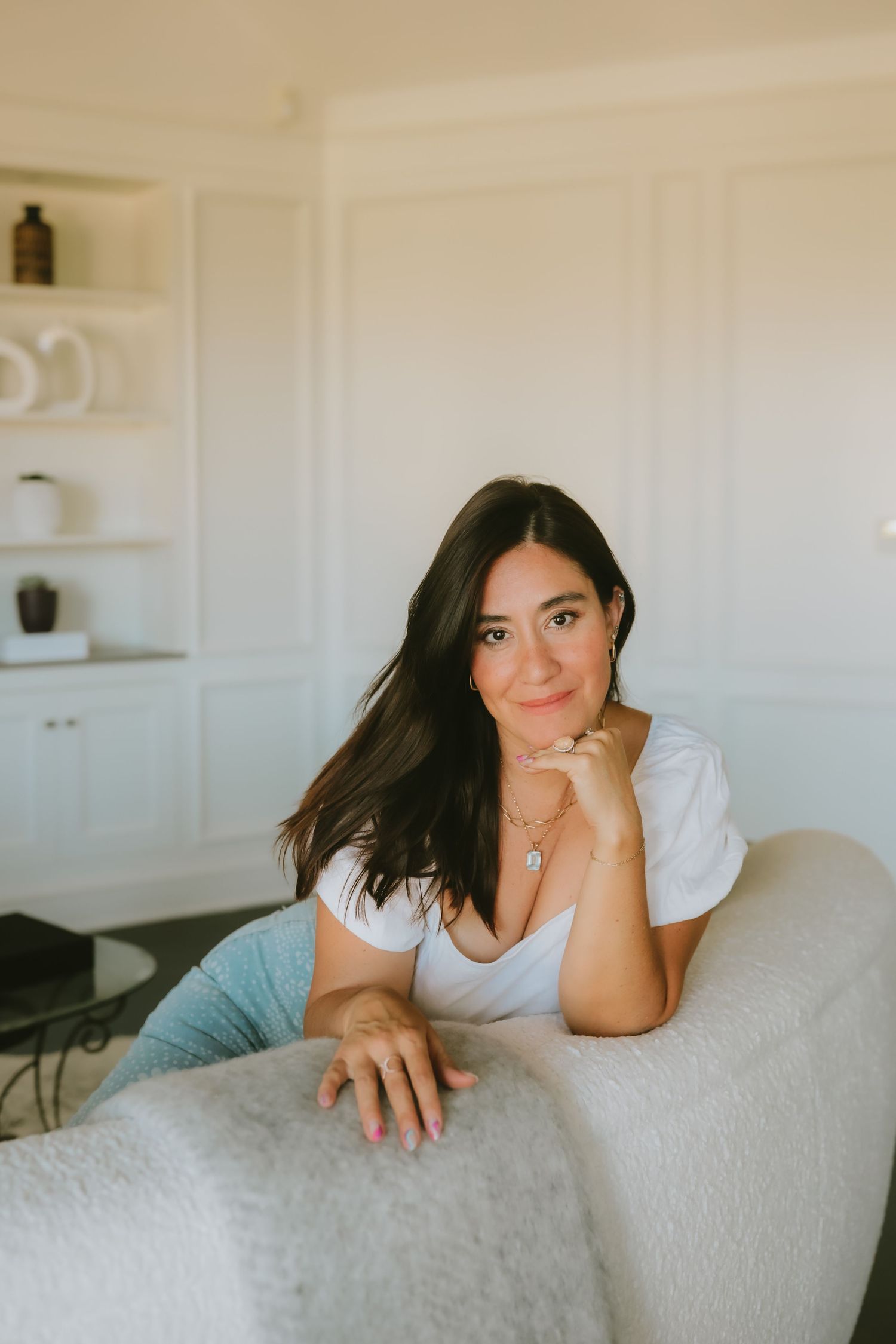 A woman in a white t-shirt relaxes on a light gray couch against white built-in shelves in a bright, airy room.