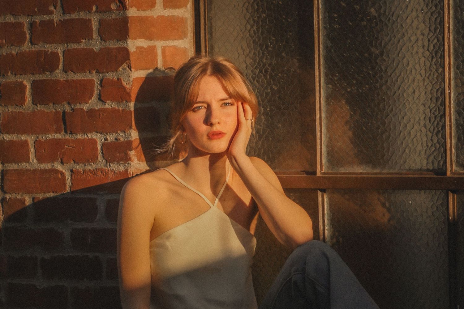 Person in white top poses near brick wall at sunset with warm golden lighting creating atmospheric mood.