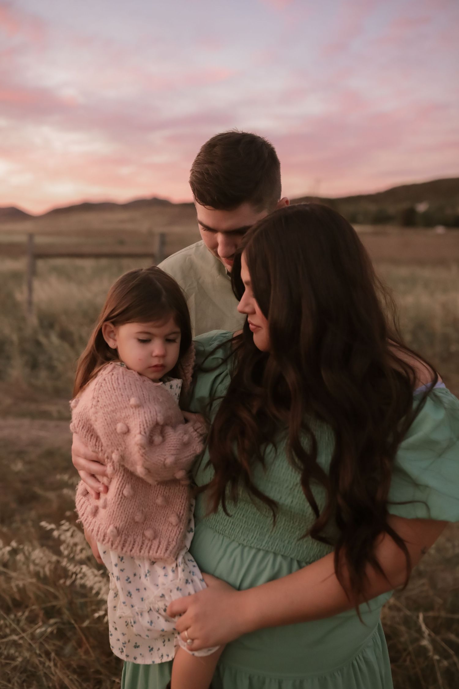 A family stands close together in a field during sunset, with a pink and orange sky in the background.