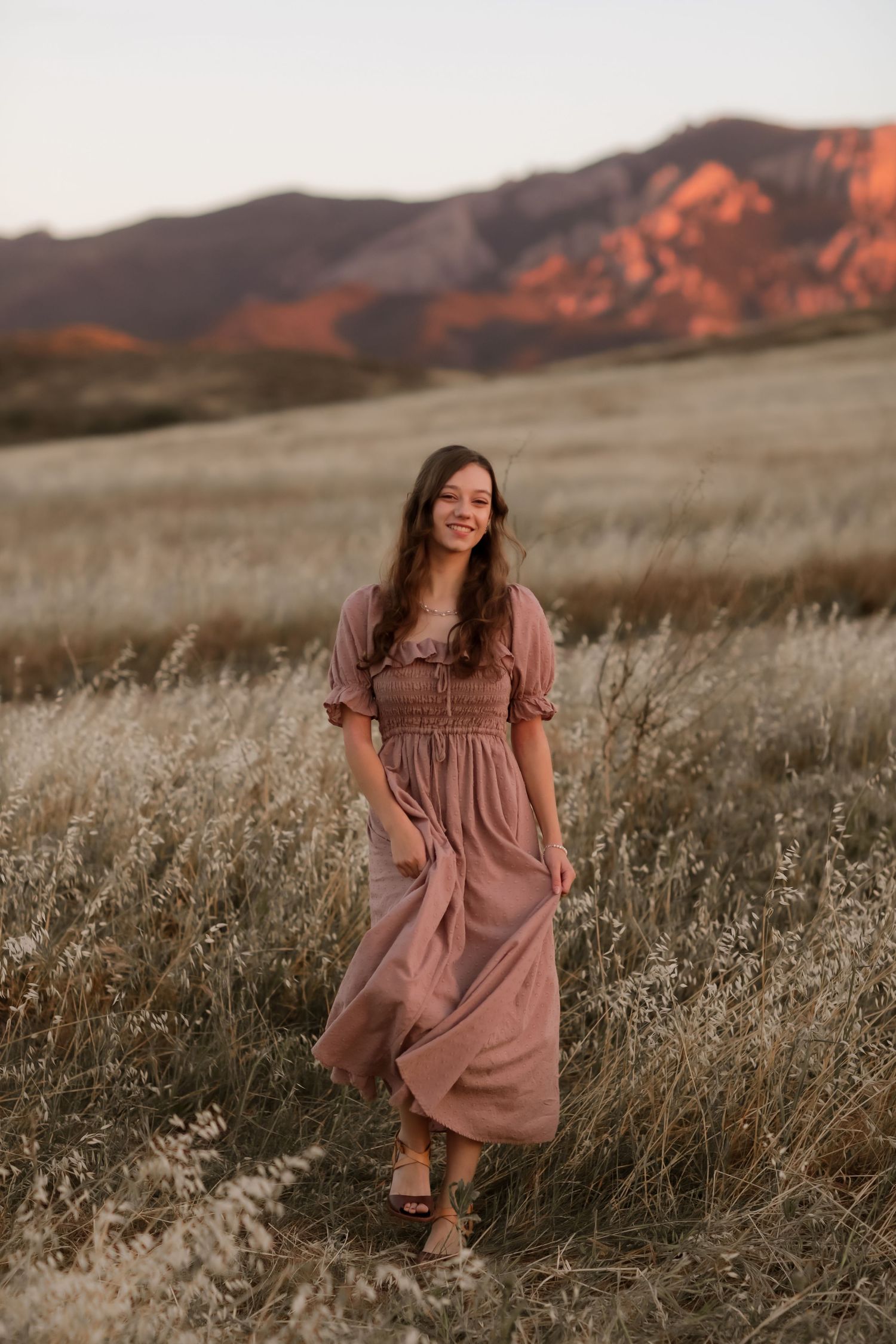 A person in a dusty rose maxi dress stands in a field of tall grass at sunset with mountains in the background.