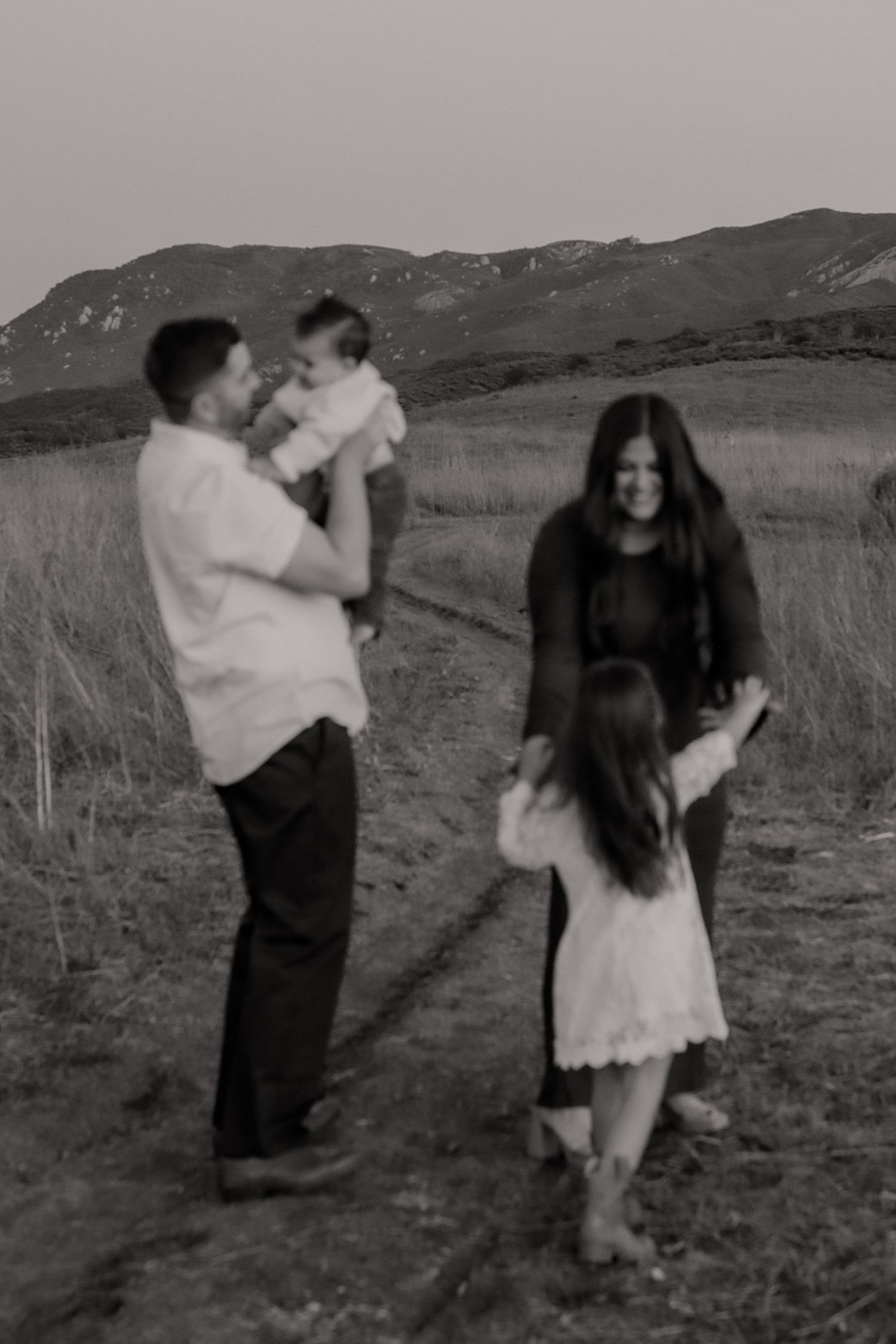 A couple dances playfully in a field with mountains visible in the background, captured in a black and white photograph.