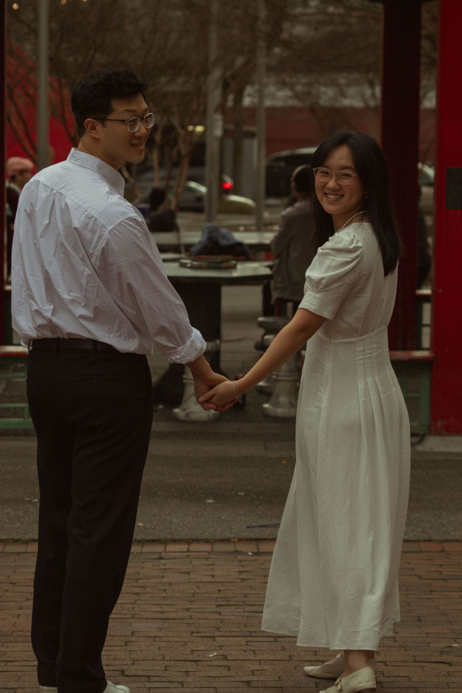 Couple holding hands outdoors, woman in white dress smiling at camera, man in white shirt and black pants, urban setting.