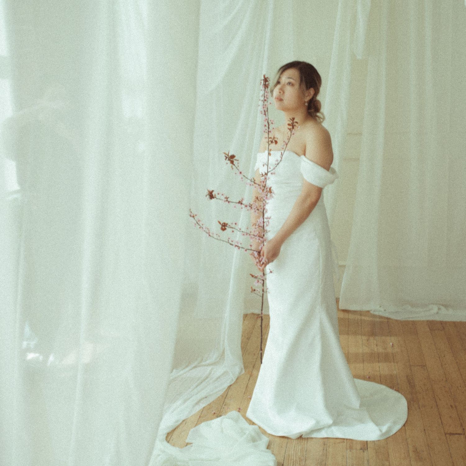 Bride in white off-shoulder gown holds cherry blossom branch, standing by sheer curtains on hardwood floor.