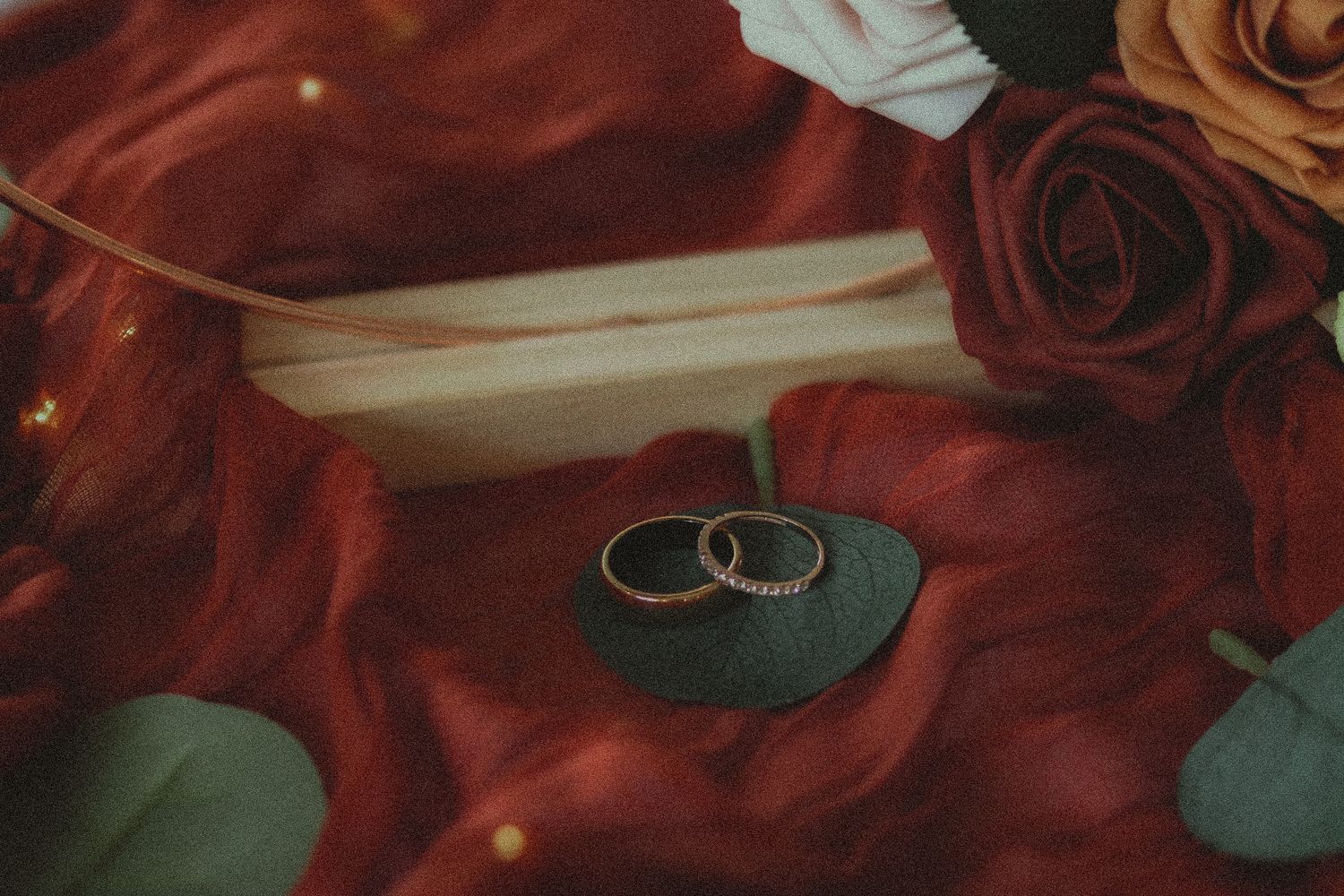 Two gold wedding rings rest on red velvet fabric beside roses and eucalyptus leaves.