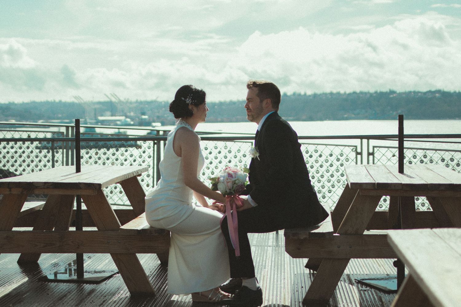 Bride and groom sitting at a waterfront picnic table on a deck, sharing a romantic moment on their wedding day.
