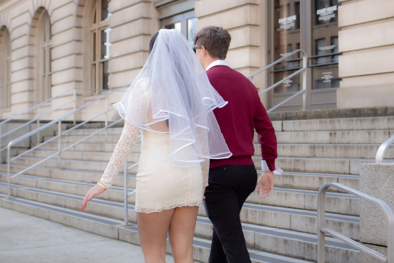 Bride in white mini dress and veil walks hand-in-hand with groom in burgundy sweater outside a stone building in Tacoma Courthouse