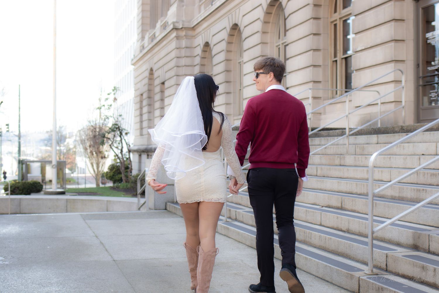 Bride in white dress and veil walks hand-in-hand with groom in red sweater outside Tacoma WA courthouse a stone building.