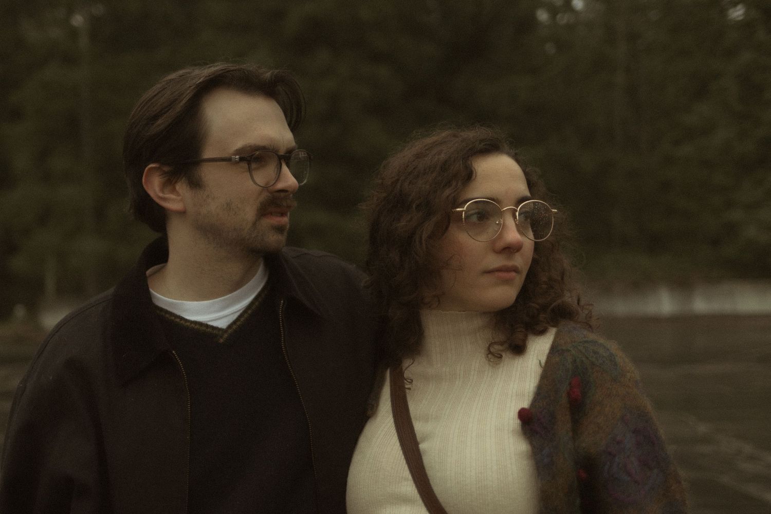 A man and woman with glasses stand close together outdoors near trees in a moody, overcast setting. Cinematic Seattle engagement photos.