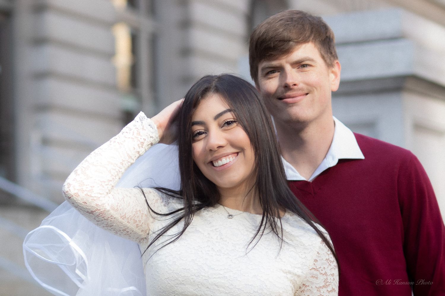 A smiling couple poses outdoors, woman in white sweater touching her hair, man in burgundy shirt behind her.