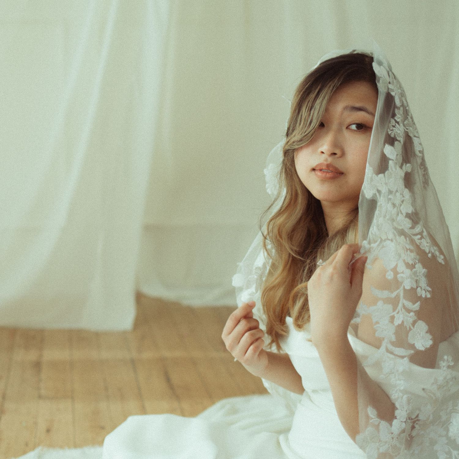 A young bride in a white lace wedding dress and floral veil sits gracefully on a wooden floor.