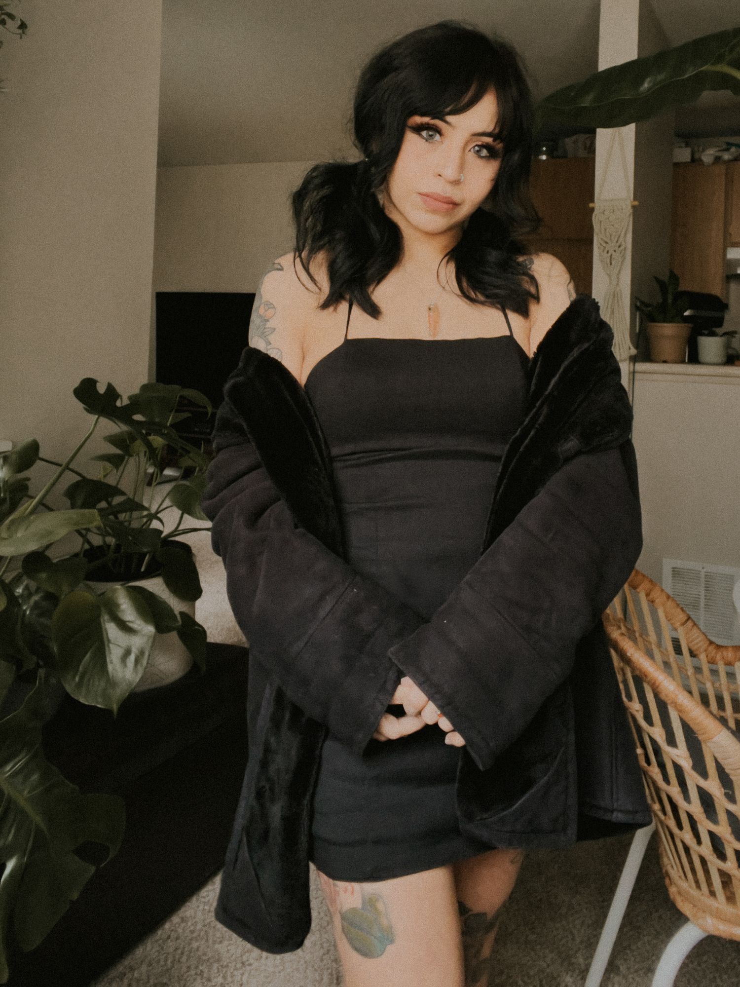 Young woman with black hair wearing a black mini dress and oversized jacket, posing indoors with tattoos visible.