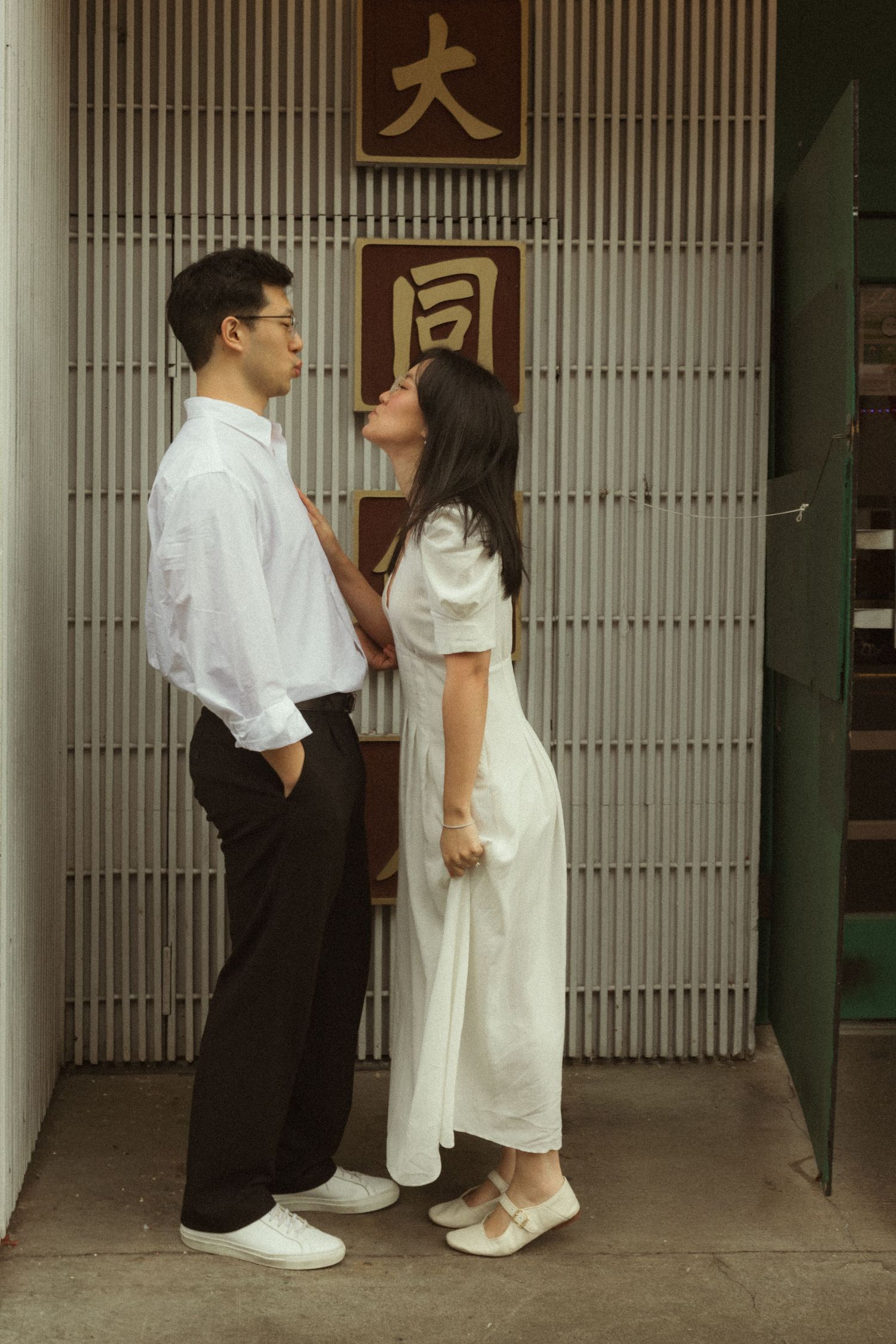 A couple in white outfits share a romantic moment near a wall with Chinese characters in an urban setting.