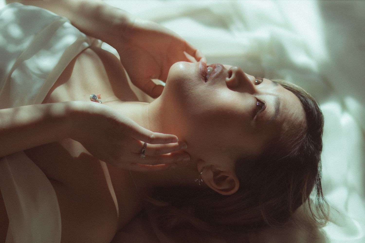 A young woman lying on white bedding, head tilted back, in soft warm light with a cinematic  dreamy, serene expression.