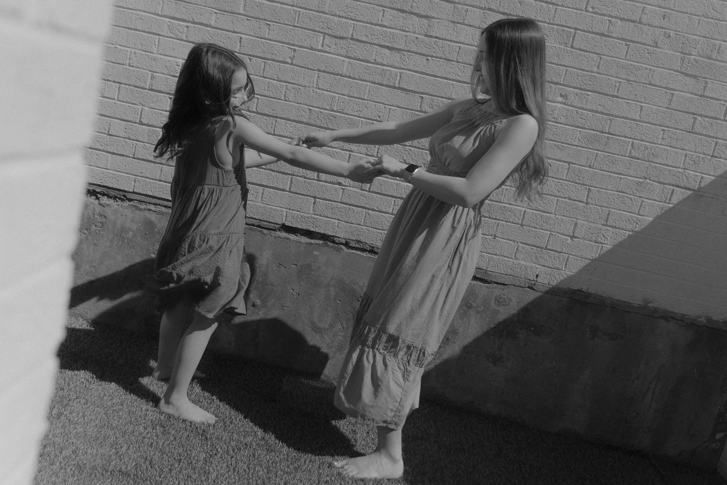 Two young girls in dresses hold hands and dance together outdoors near a brick wall in black and white photo.