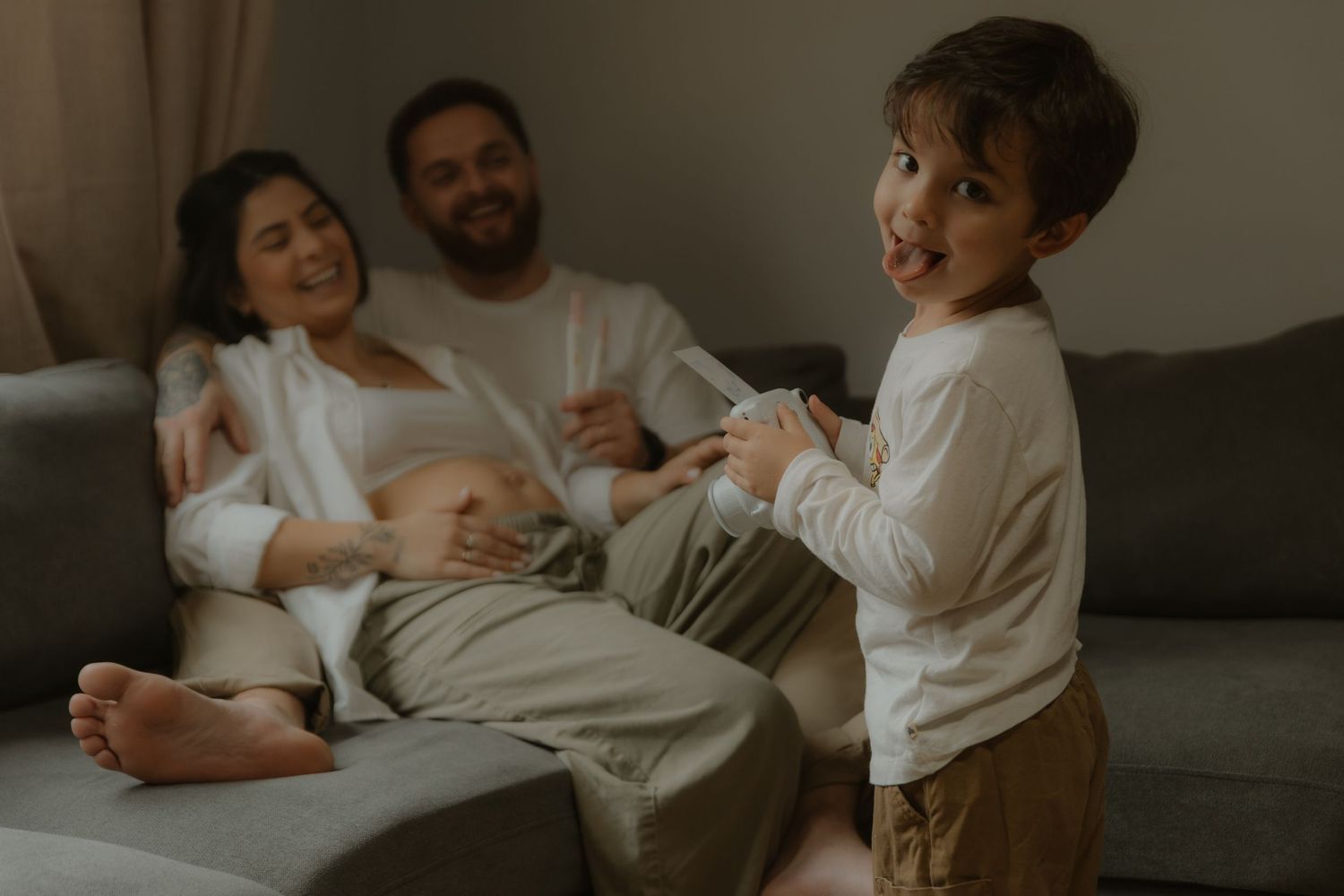 Pregnant woman relaxing on sofa with husband while young toddler boy smiles and plays in cozy living room.