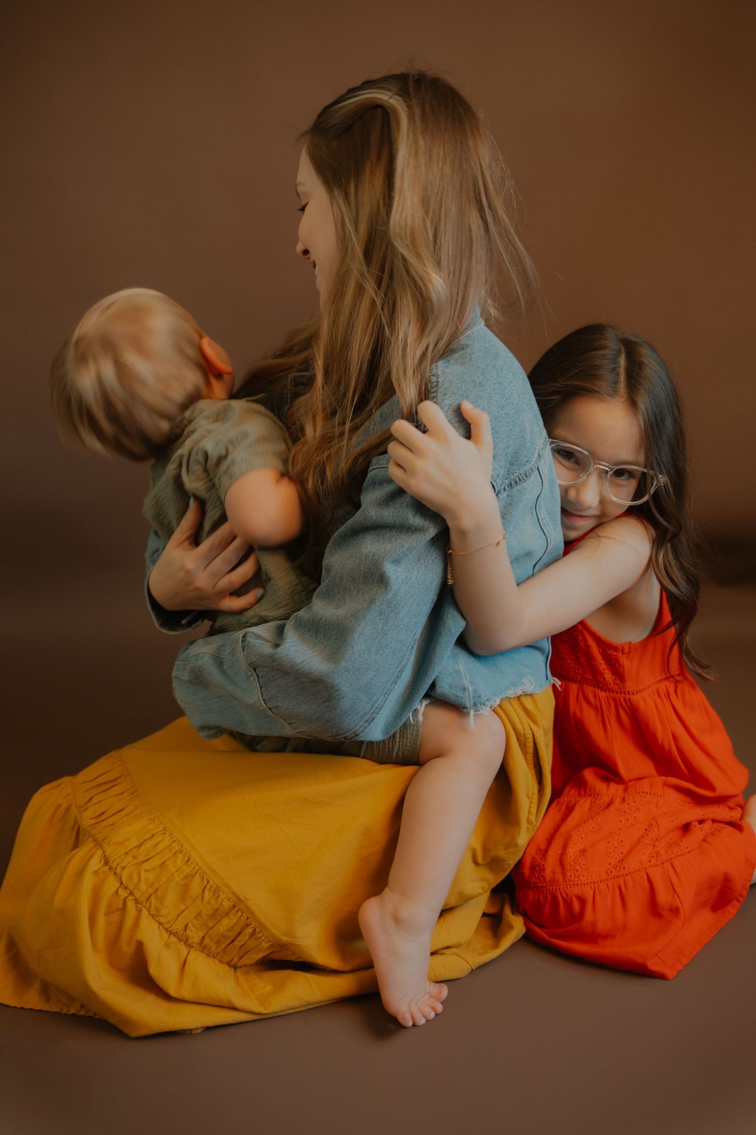 A mother sits on the floor hugging two young children, a toddler and a girl in glasses wearing a red dress.