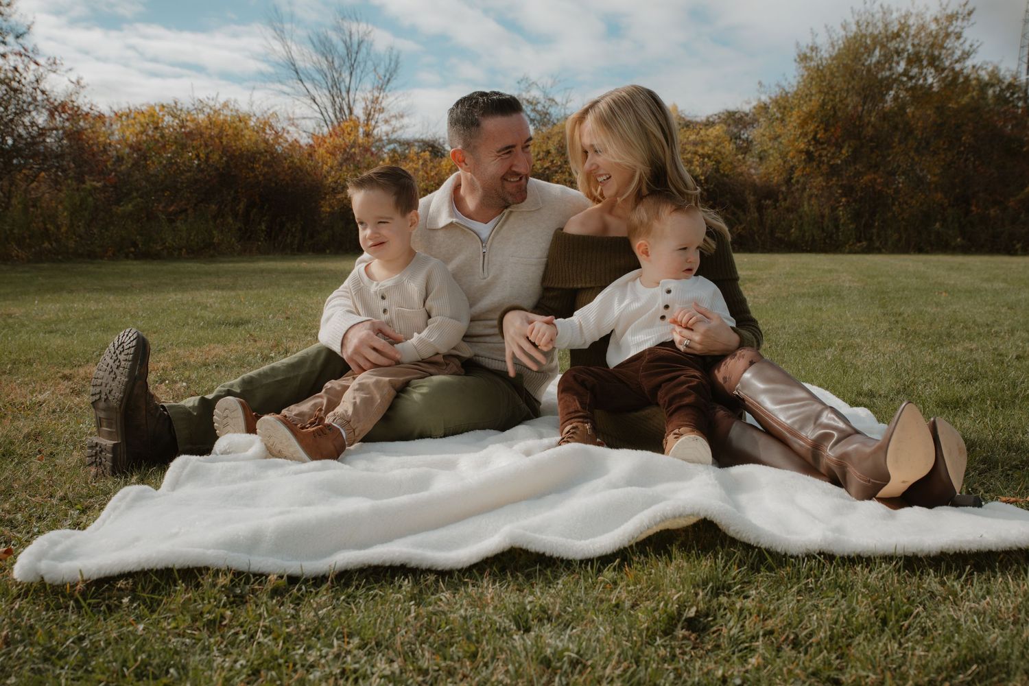 Family of four sitting on white blanket in autumn field, parents with two young boys in coordinated earth-tone outfits.