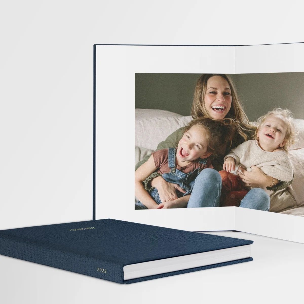 Navy hardcover photo book open to a joyful family portrait of a mother laughing with two young children on a couch.