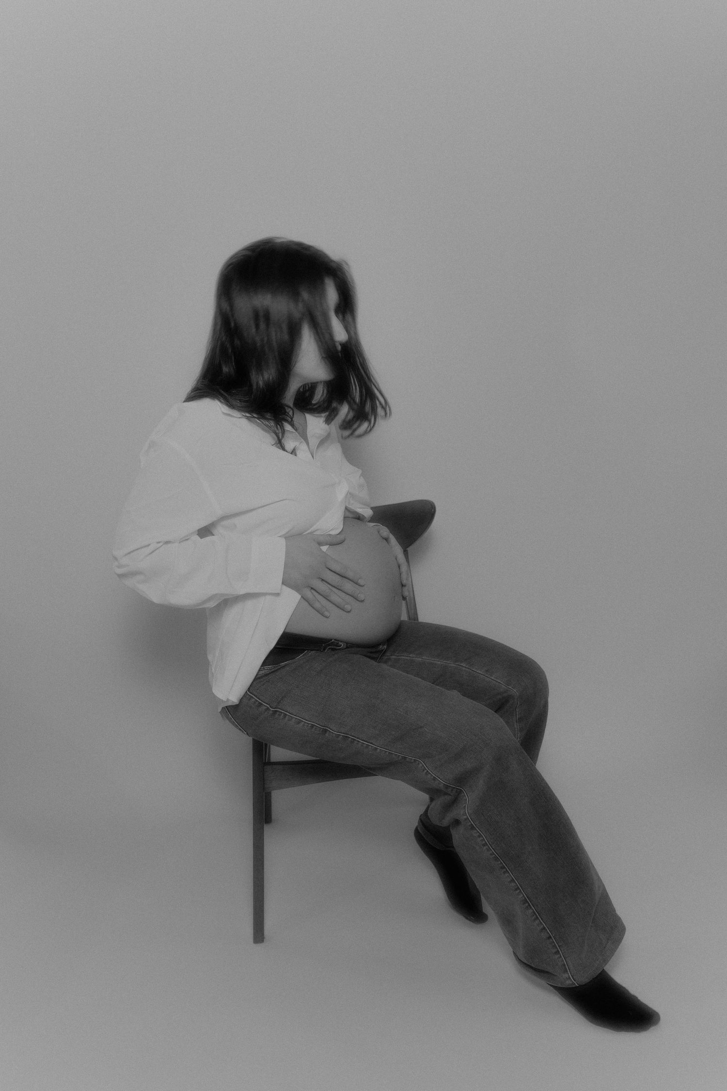 Black and white photo of a pregnant woman sitting on a chair, cradling her belly, wearing a white shirt and jeans.