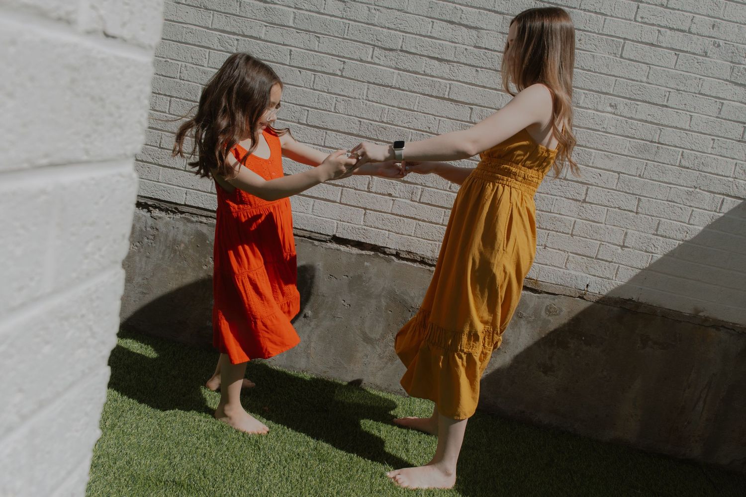 Two barefoot girls in colorful red and orange dresses hold hands and dance together on green grass by a brick wall.
