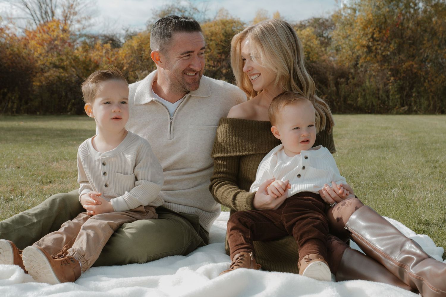 Happy family of four sitting on a blanket in a green field during autumn, smiling together outdoors.