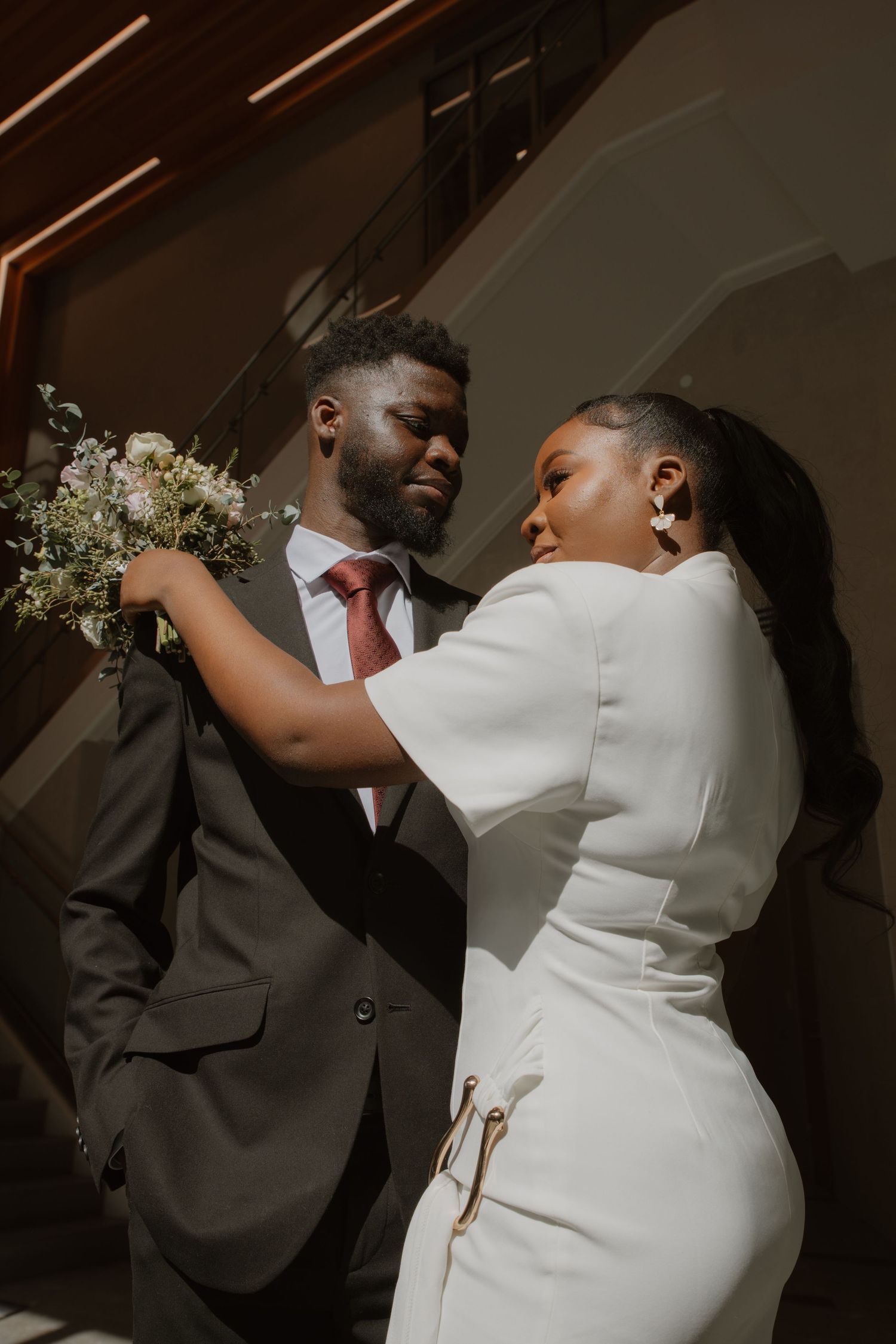 A couple in formal attire embraces while holding a white floral bouquet.