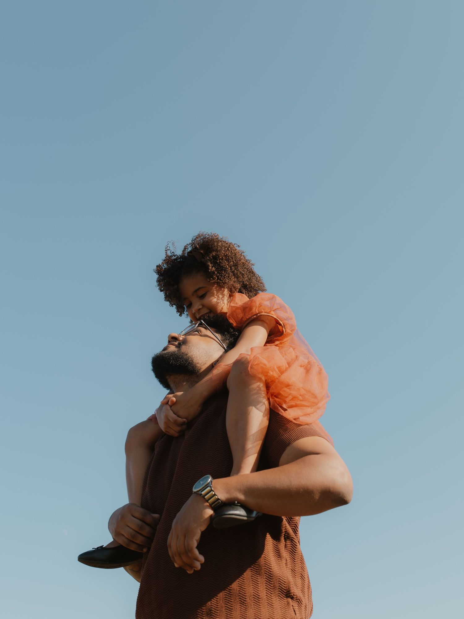 A man lifts a curly-haired girl in an orange dress against a clear blue sky.