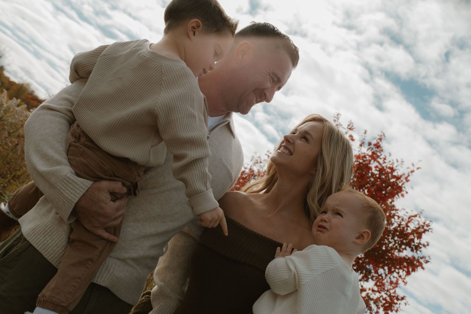 Happy family of four enjoying an autumn outdoor photo shoot under a cloudy sky with colorful fall foliage.