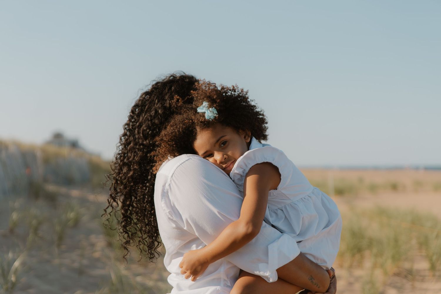 A tender embrace between two figures on a sunny beach with natural grasses in the background.