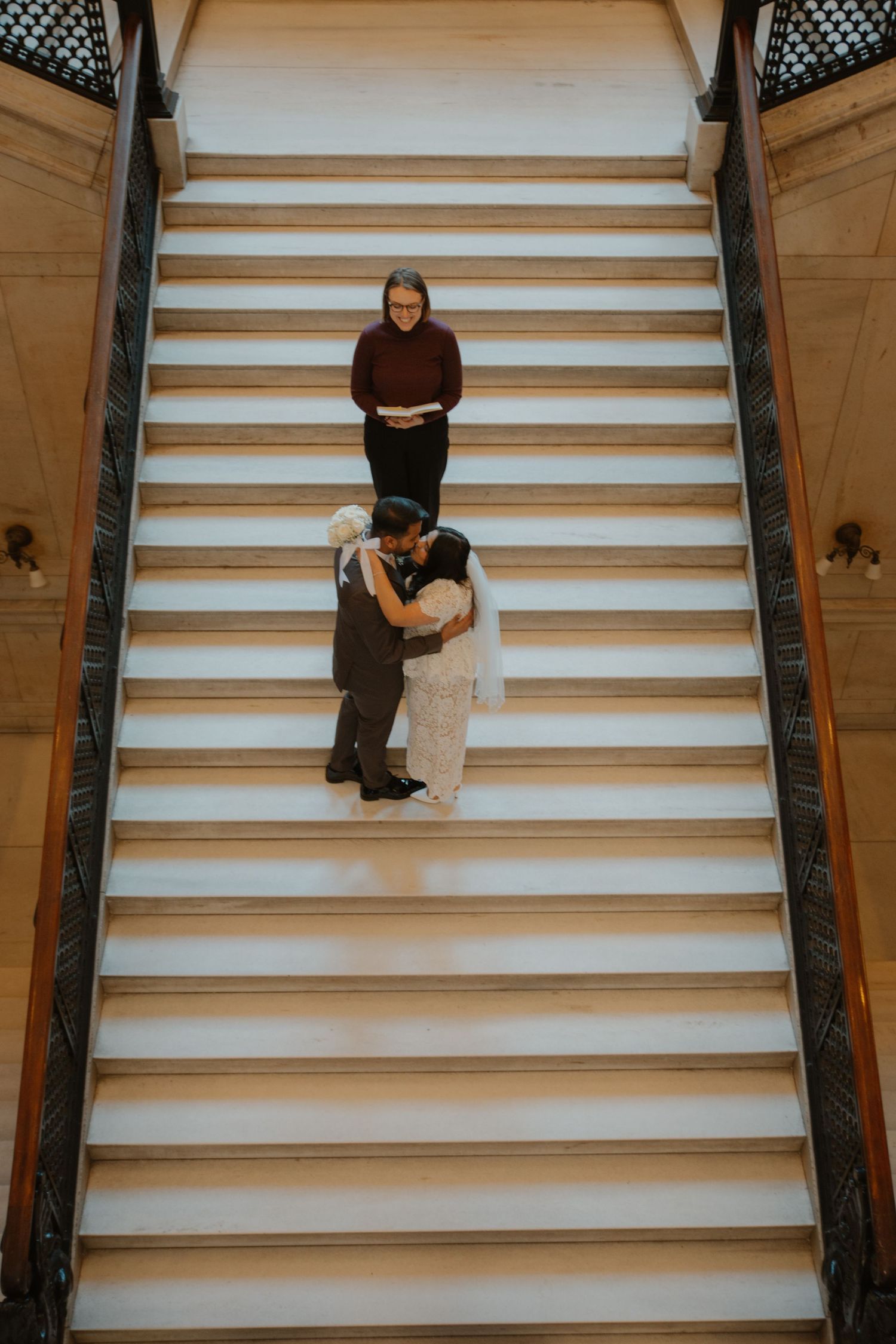 Couple shares first kiss on grand staircase during intimate wedding ceremony with officiant standing behind them.