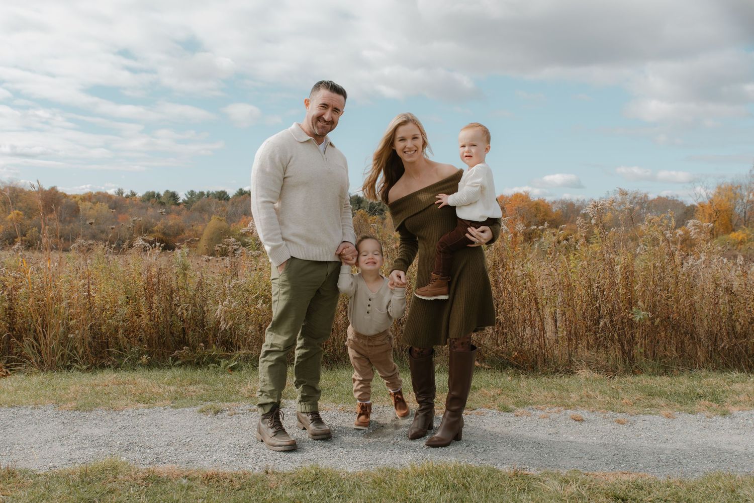 Family of four posing outdoors in autumn, parents holding young children on a scenic fall path with colorful foliage.