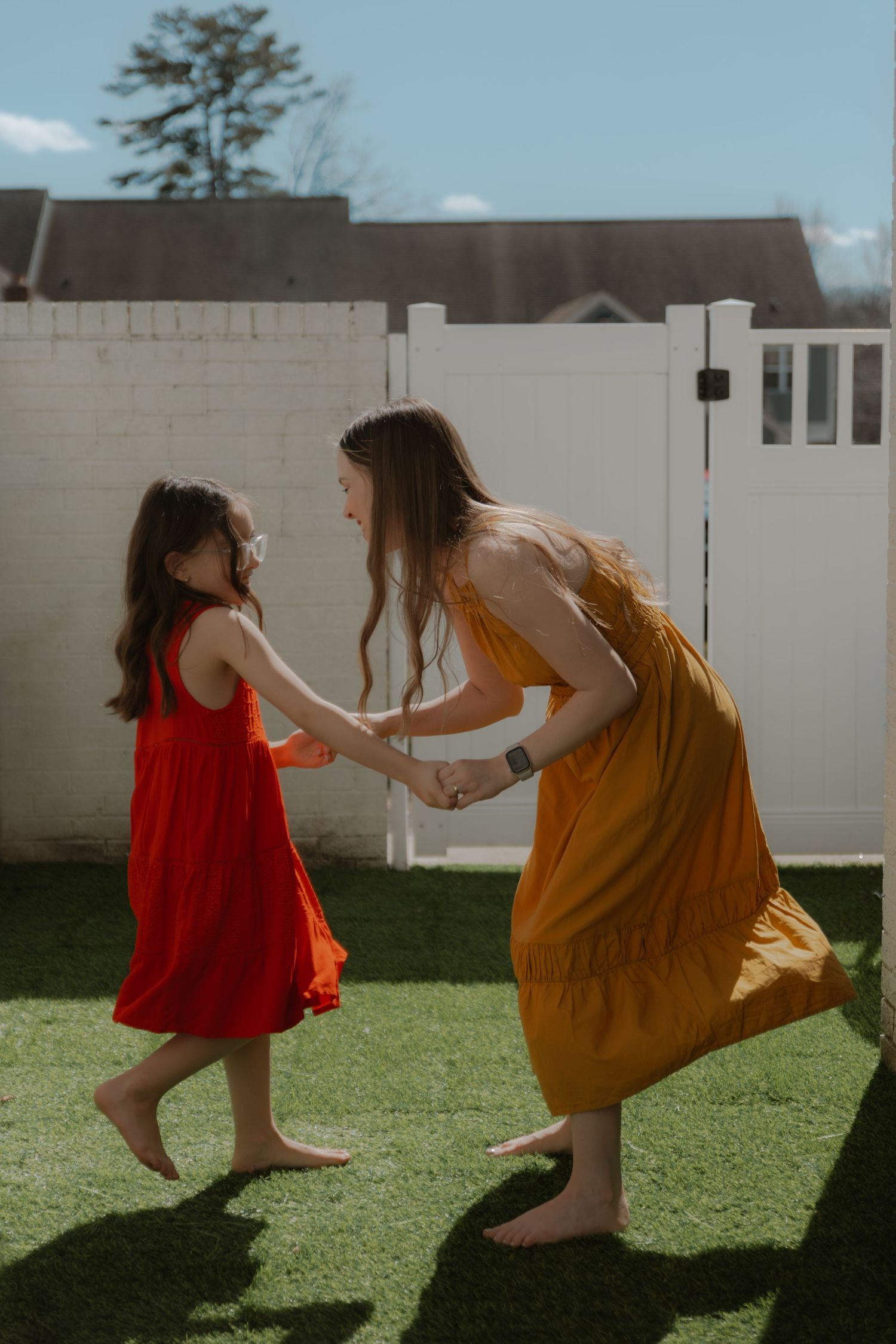 A mother and daughter in colorful dresses hold hands and dance barefoot on green grass in a sunny backyard.