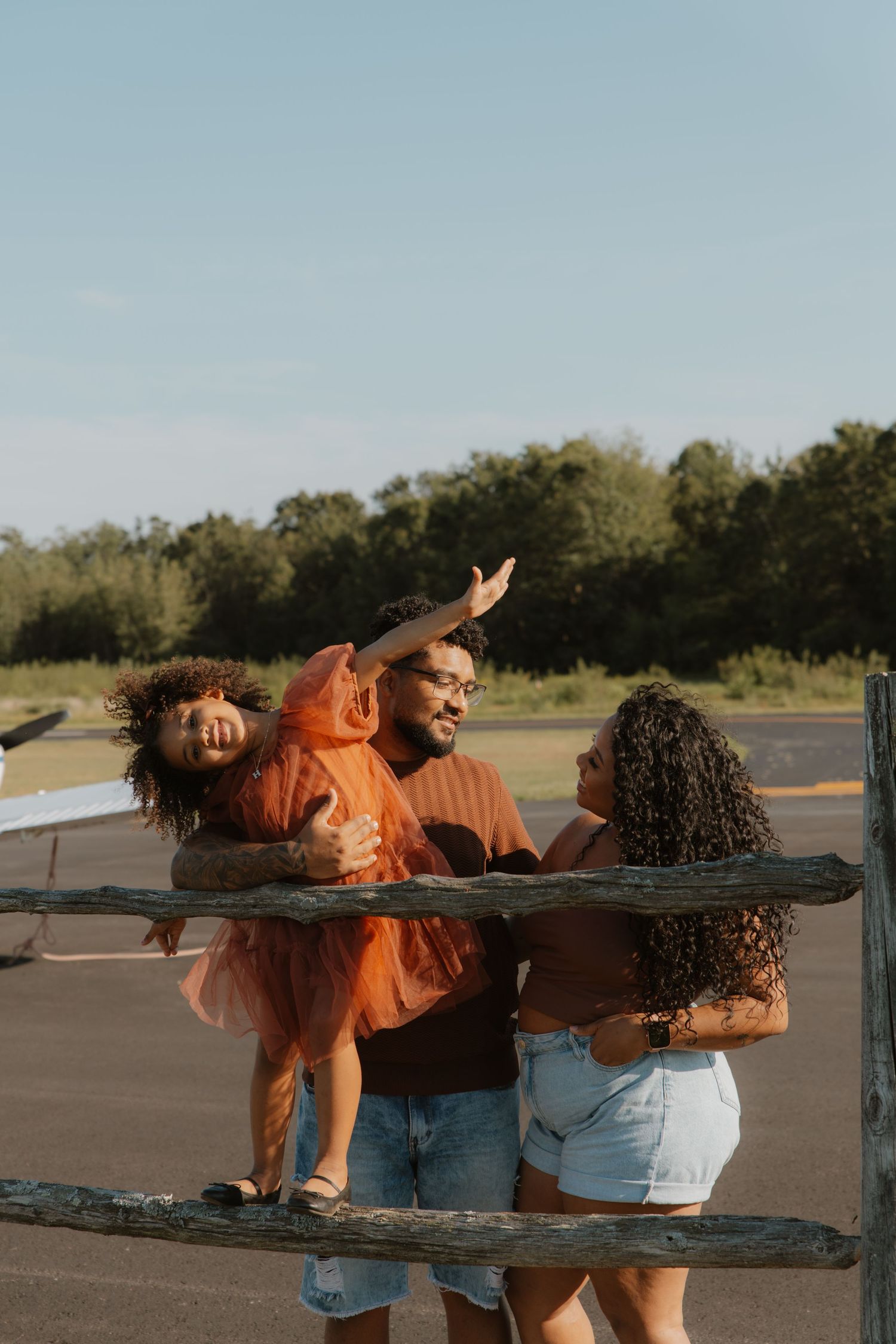 A joyful family of three poses outdoors near a wooden fence, with a man holding a laughing girl in an orange tutu.