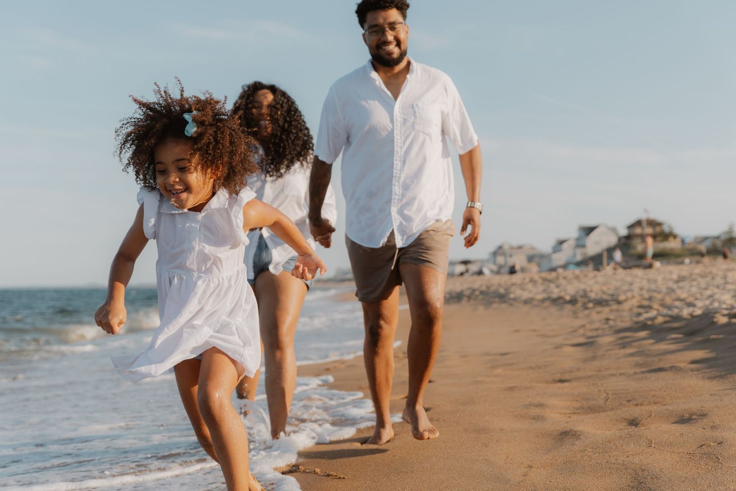Happy family running on a sunny beach, parents and young girl in white outfits enjoying a seaside vacation.