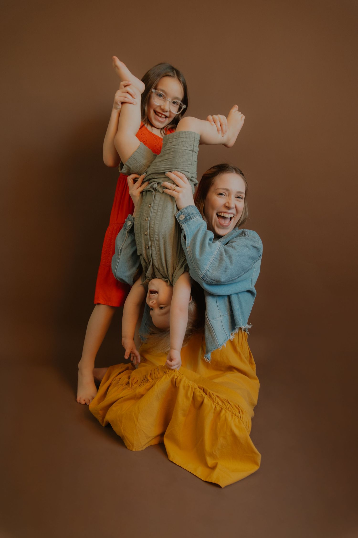 Three playful girls posing together in a studio, wearing colorful outfits against a brown background.