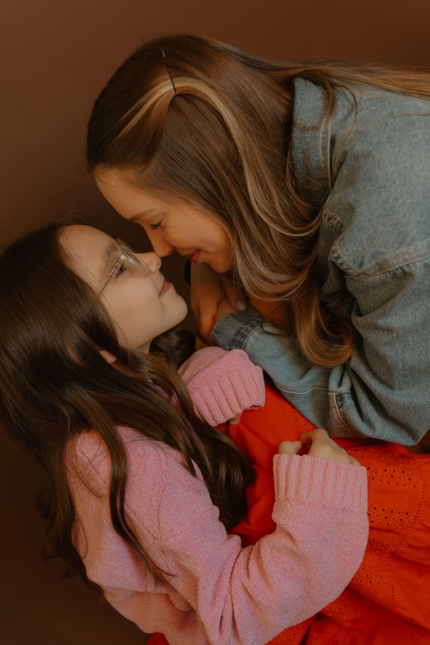 A woman and young girl with long brown hair share a tender nose-to-nose moment, wearing pink and denim outfits.