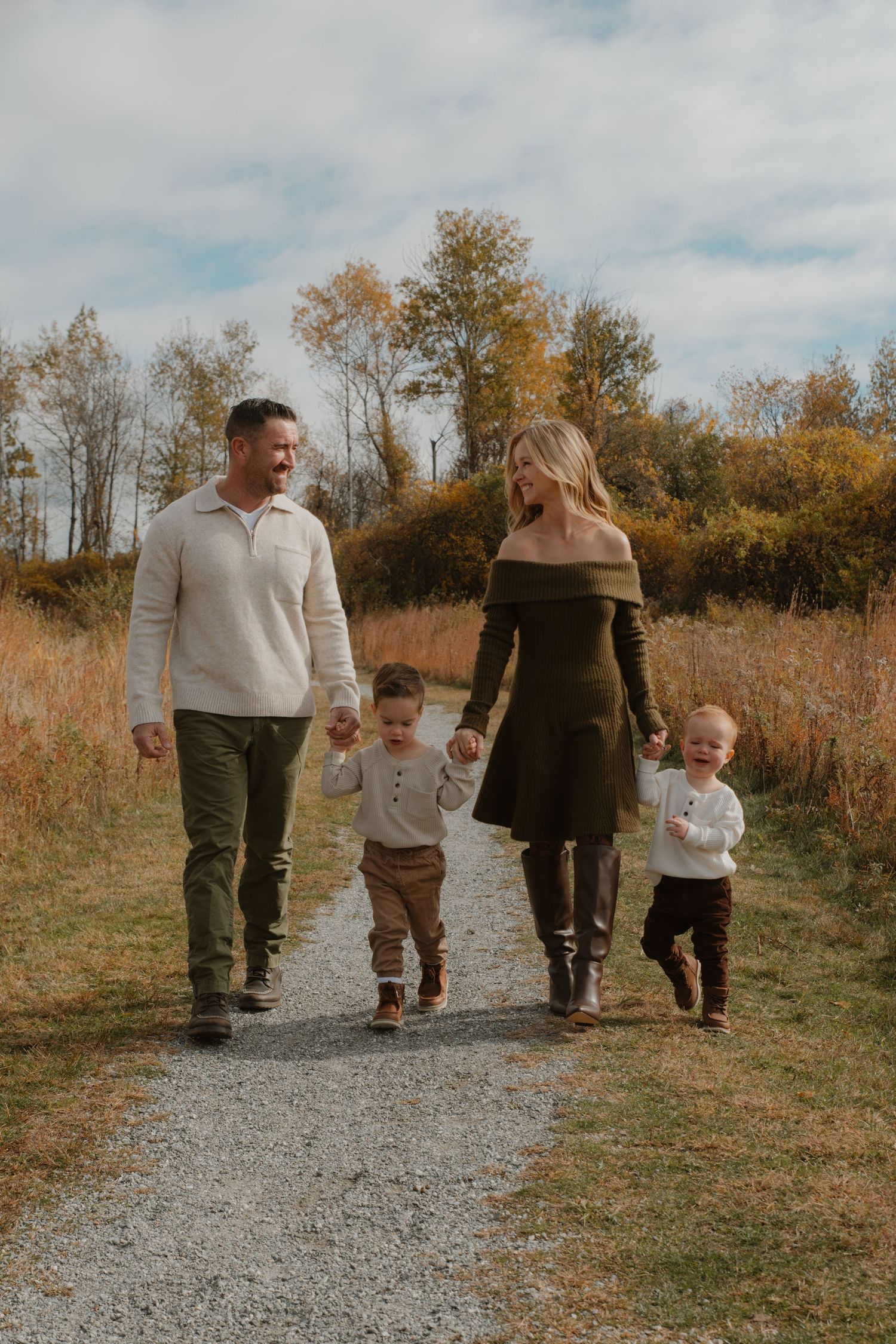 Family of four walking on a gravel path in autumn, parents holding hands with two toddlers in a scenic fall landscape.