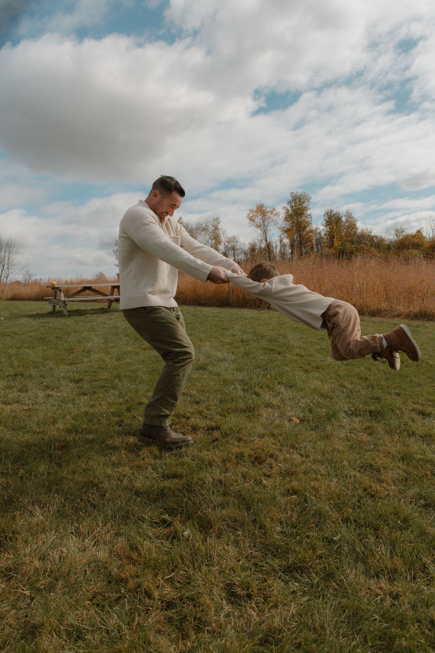 A father swings his young child by the arms in a grassy field on a cloudy autumn day.
