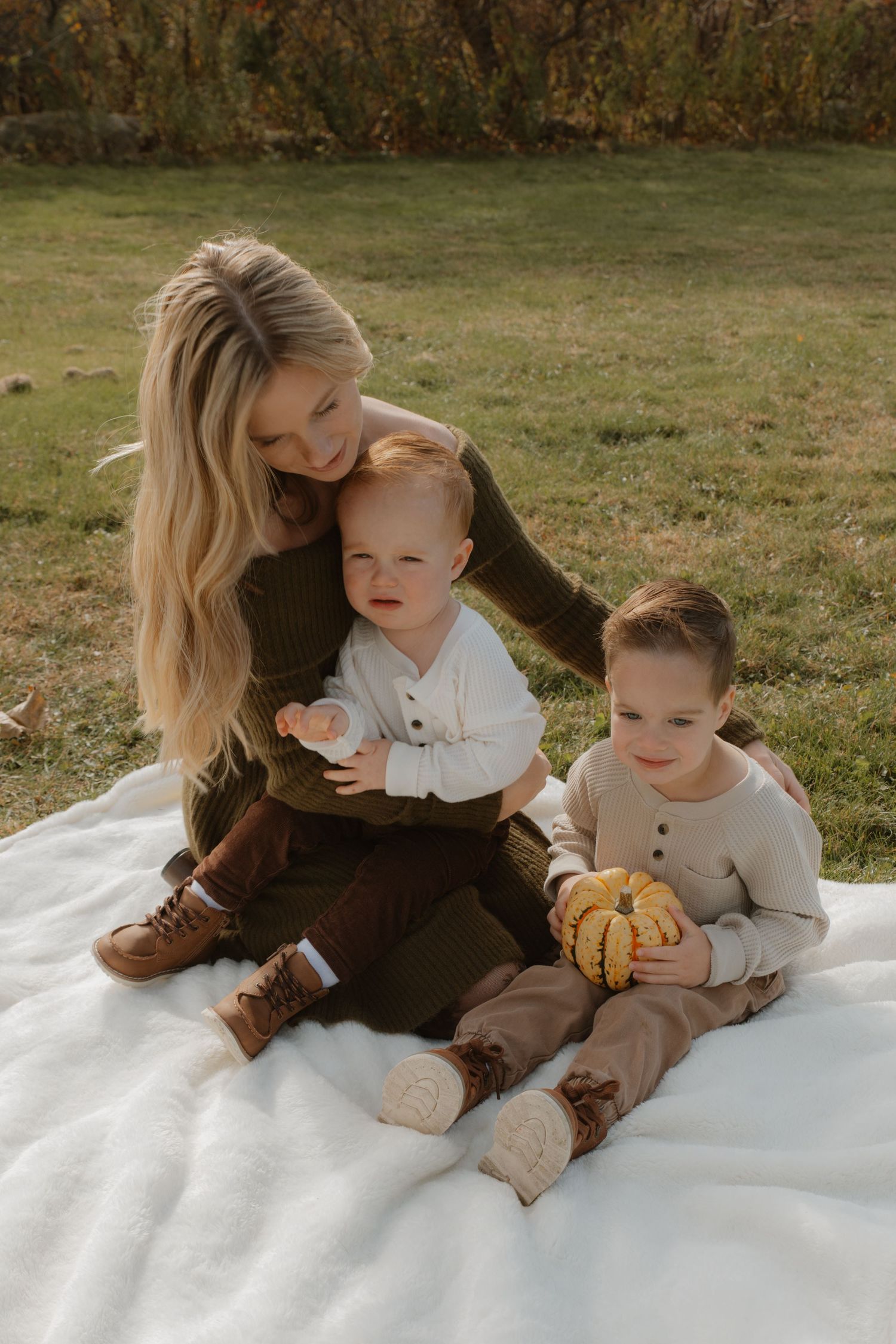 A mother sits on a white blanket outdoors with her two young children, holding a small pumpkin in autumn.