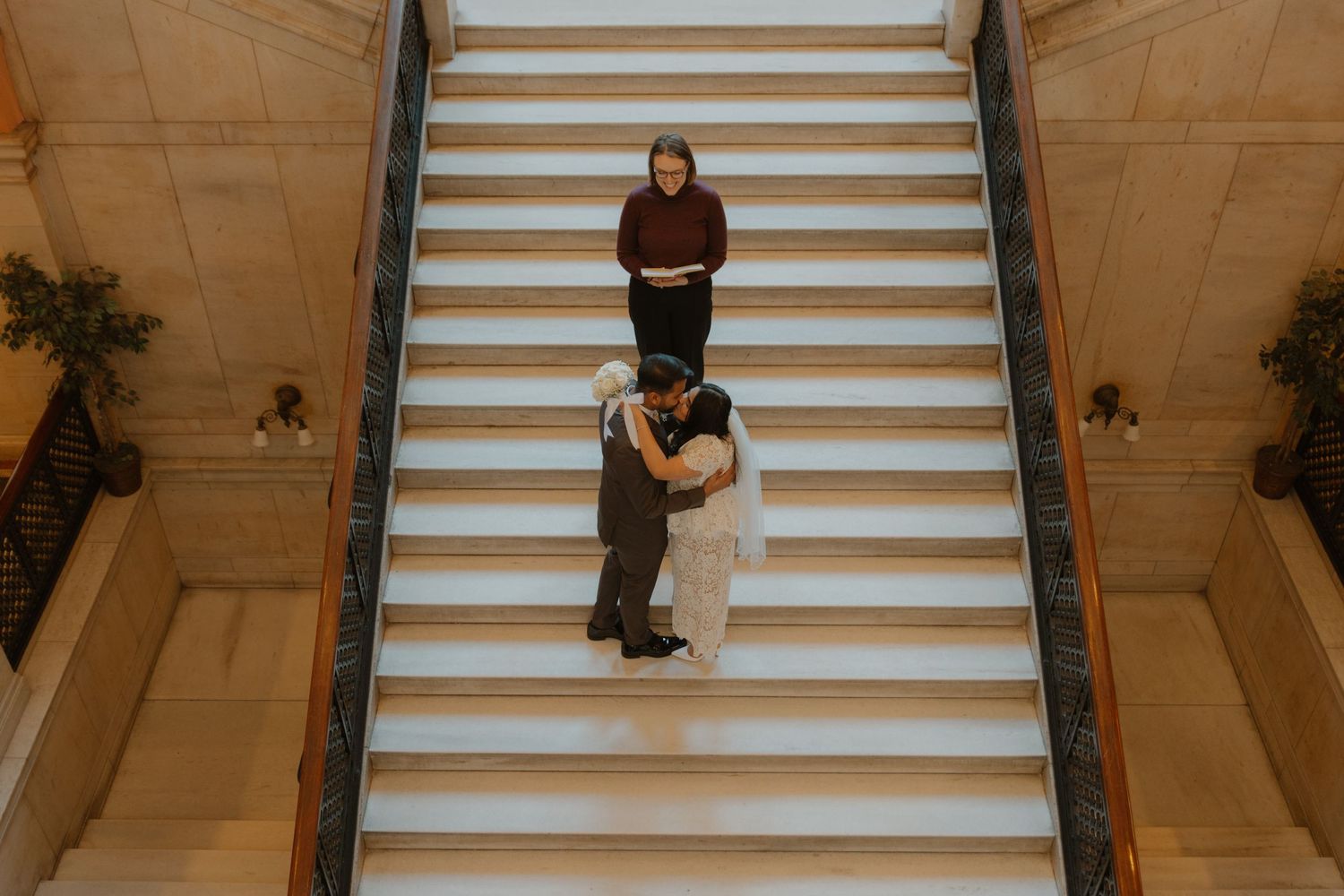 Two people pose on white marble stairs in a grand interior setting.