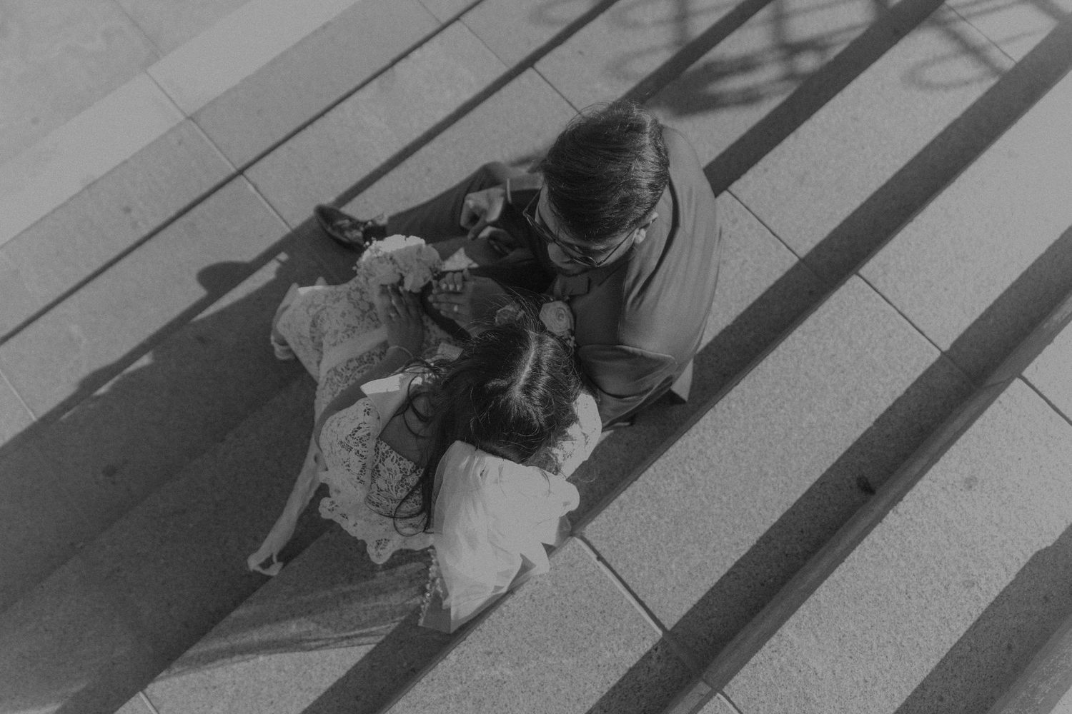 Black and white aerial view of two people sitting together on sunlit steps, sharing a tender moment.